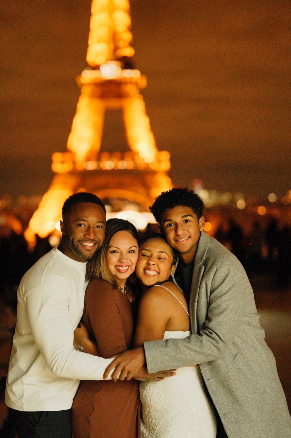 Bill Braxton with his wife and two children embracing in front of the Eiffel Tower glowing gold at night